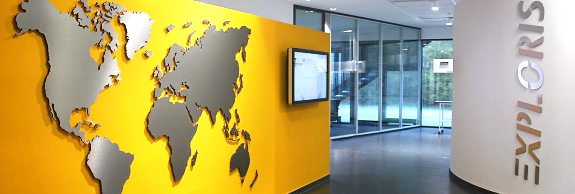 Innenbereich mit gelber Wand und silberner Weltkarte, daneben ein Monitor; rechts eine weiße, gebogene Wand mit dem Schriftzug „EXPLORIS“ in großen Buchstaben.